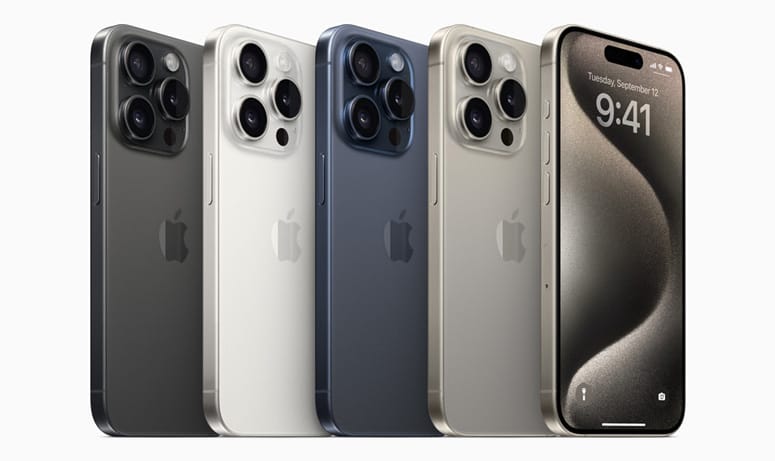 Quatro smartphones Apple iPhone 15 alinhados lado a lado nas cores Titânio Preto, Branco, Azul e Natural. O aparelho da extrema direita exibe a tela frontal acesa com o papel de parede padrão.